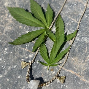 May include: A handmade necklace featuring a clear quartz crystal pendant, brass-colored beads, and decorative triangle charms. The necklace is displayed with green cannabis leaves on a textured gray surface.