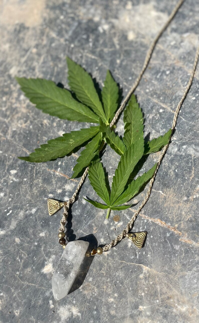 May include: A handmade necklace featuring a clear quartz crystal pendant, brass-colored beads, and decorative triangle charms. The necklace is displayed with green cannabis leaves on a textured gray surface.