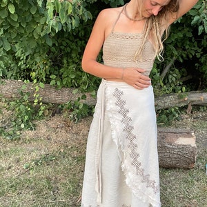 Könnte beinhalten: Ein beiges Häkeltop und ein cremefarbener Wickelrock mit geometrischem Stickmuster. Der Rock hat einen Fransenrand und eine lange Bindung. Das Outfit wird im Freien getragen.