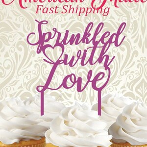 May include: Four cupcakes with white frosting are displayed. The cupcakes sit on a black and white patterned ribbon. The image has text that reads "American Made", "Sprinkled with Love", and "A Part of the Country Road Family of Shops".
