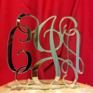 May include: A silver mirrored cake topper with a cursive monogram of the letters "C, G, and P". The topper is on a white cake.