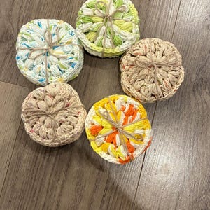 May include: Six handmade crochet coasters in a variety of colours. The round coasters feature a textured, floral design. Colours include white, green, yellow, orange, and beige. Each coaster is tied with twine.