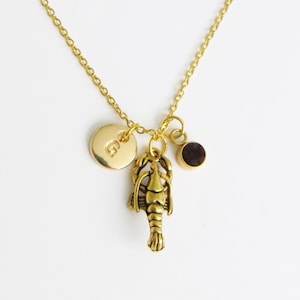 May include: Gold necklace with a lobster charm, a small round charm with the letter 'G' and a small round purple gemstone.