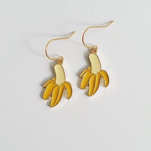Puede incluir: Un par de pendientes de tono dorado con un diseño de plátano amarillo. El plátano se representa con una cáscara blanca y una pulpa amarilla.