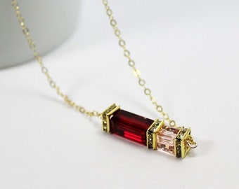 May include: A gold necklace with a pendant. The pendant has a rectangular, deep red gemstone and a smaller, light pink gemstone. The gemstones are set in a gold frame. The necklace is displayed on a white surface.