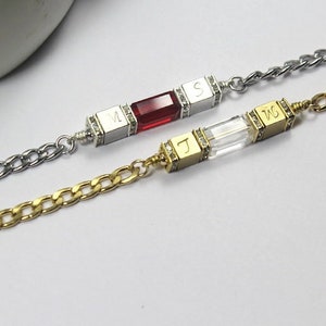 May include: Two silver and gold chain bracelets with personalized charms. The silver chain bracelet has a red gemstone and the letters "M" and "S" engraved on the charms. The gold chain bracelet has a clear crystal and the letters "J" and "W" engraved on the charms.