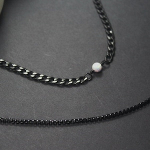 May include: Two black chain necklaces, one thicker than the other, with a small white bead on the thicker chain.