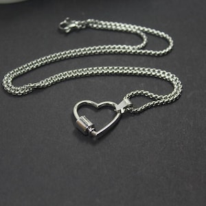 May include: A silver chain necklace with a heart-shaped pendant. The pendant is open and has a small cylinder in the center.