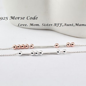 May include: Two silver chains with a bar and beads. The top chain has five rose gold beads and the bottom chain has four silver beads. The text "925 Morse Code Love. Mom. Sister. BFF, Aunt, Mama." is above the chains.