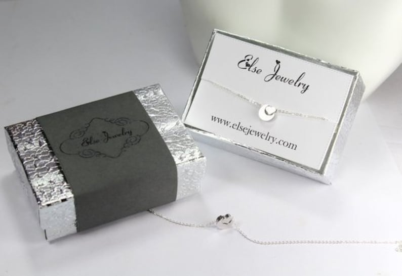 May include: A silver necklace with a small circle pendant, packaged in a grey box with the text "Else Jewelry" and a white box with the text "Else Jewelry" and the website address "www.elsejewelry.com".