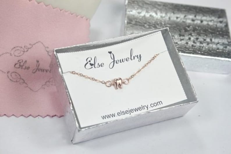 May include: A silver jewelry box with a white card inside. The card has the text "Else Jewelry" and the website address "www.elsejewelry.com". A delicate gold chain necklace with a small tooth charm is displayed on the card.