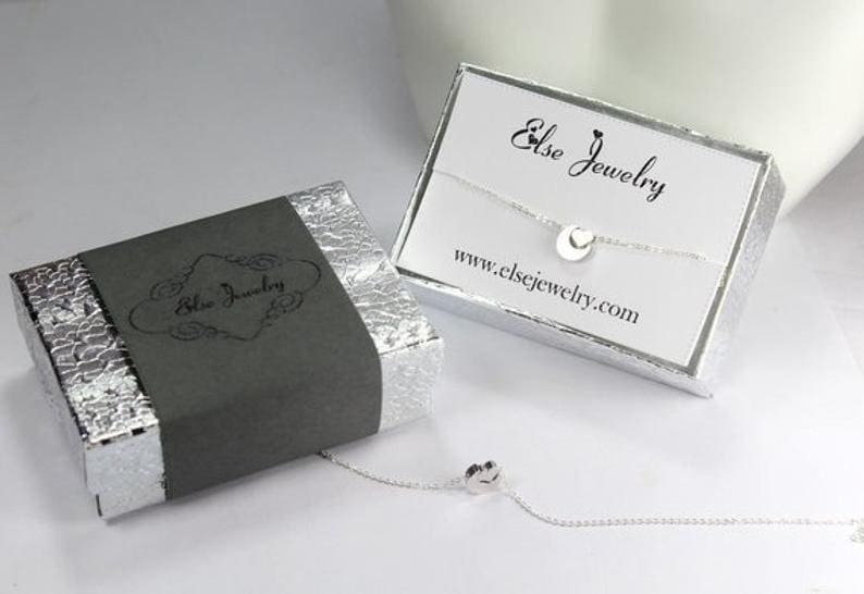 May include: A silver necklace with a small heart charm comes in a gray and silver box with the text "Else Jewelry" on the box and the website address "www.elsejewelry.com" on the white card inside the box.