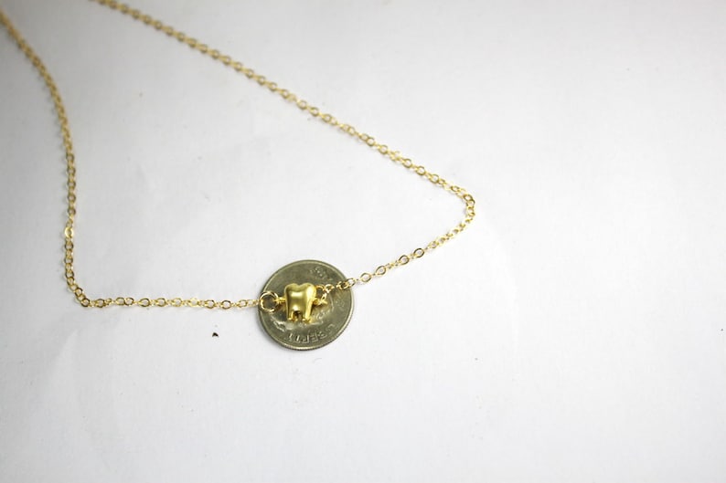 May include: A gold chain necklace with a small gold tooth charm. The charm is hanging from a coin.