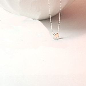 May include: A fine silver necklace with a small, circular pendant. The pendant has a silver outer ring and a gold-coloured centre. The necklace is presented on a white surface, creating a minimalist aesthetic.
