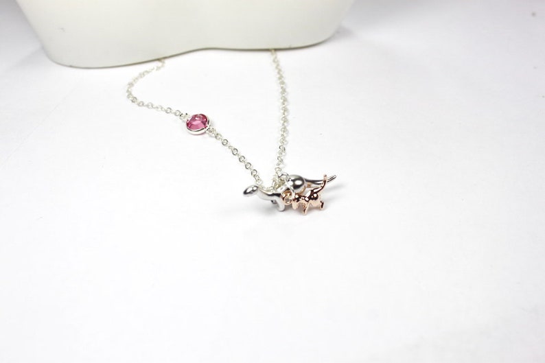 May include: A silver chain necklace with a pink gemstone charm and a silver and rose gold dinosaur charm.