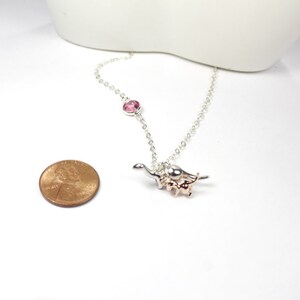 May include: A silver chain necklace with a pink crystal charm and a silver and rose gold dinosaur charm. The necklace is about the size of a UK penny.