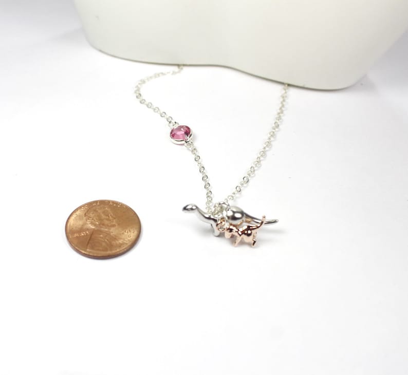 May include: A silver chain necklace with a pink crystal charm and a silver and rose gold dinosaur charm. The necklace is about the size of a UK penny.