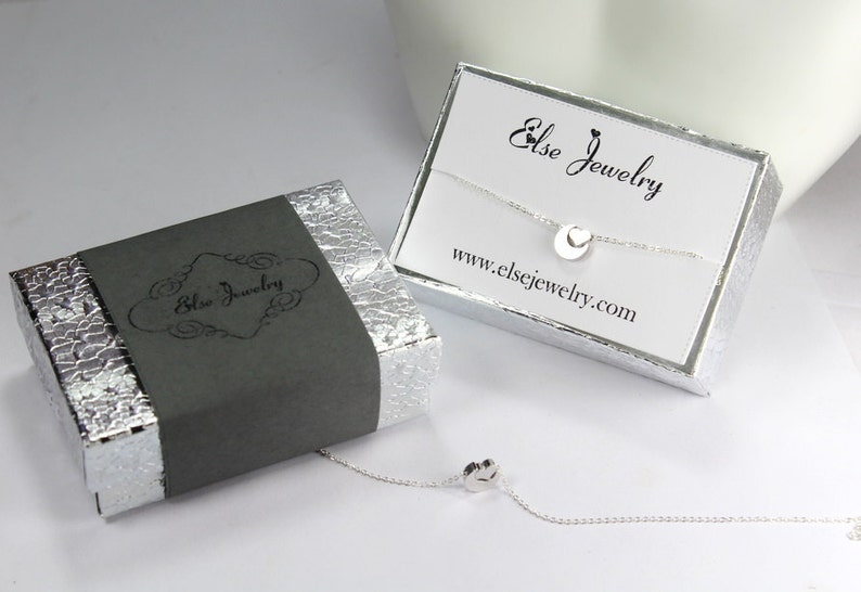 May include: A silver necklace with a small circle pendant featuring a heart cutout. The necklace is displayed in a silver box with a black label that reads "Else Jewelry" and a white card with the text "Else Jewelry" and "www.elsejewelry.com".