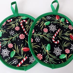 May include: Two round Christmas potholders with a black background featuring candy canes, snowflakes, and Christmas lights. The potholders have a green trim and a green loop for hanging.