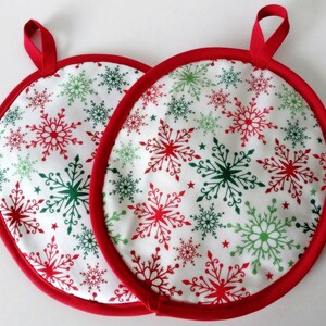 May include: Two round Christmas potholders with a white background, red trim, and red and green snowflake patterns. Each potholder has a red loop for hanging.