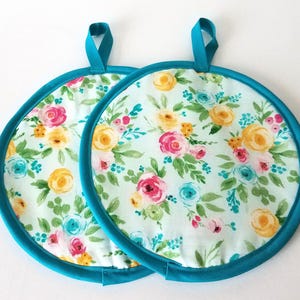 May include: Two round fabric pot holders featuring a floral pattern with yellow, pink, and teal flowers on a white background. Each pot holder has a solid teal fabric border and a matching teal fabric loop at the top for convenient hanging storage.