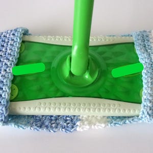 May include: A green plastic mop head attached to a green handle, featuring a removable, washable crochet cover. The cover is crafted from variegated yarn in shades of light blue, white, and dark blue, wrapping around the rectangular mop base.