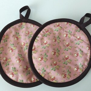 May include: Two round pink floral print pot holders with brown trim and a brown loop for hanging. The floral pattern includes red, yellow, and green flowers and leaves. The pot holders are made of fabric.