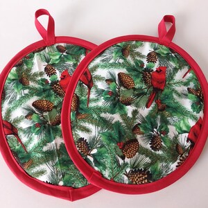 May include: Two round pot holders with red trim and hanging loops. The pot holders feature a white background with a pattern of green pine needles, brown pine cones, and red cardinals.