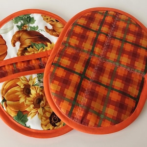 May include: Two orange and white oven mitts. One mitt features a fall harvest design with pumpkins, sunflowers, and a cornucopia. The other mitt has an orange and brown plaid pattern. Both mitts have orange trim and hanging loops.