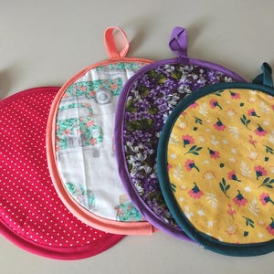 May include: Four circular fabric pot holders with hanging loops. One is pink with white polka dots, one features a white background with a teal vintage truck print, one has a purple floral pattern, and one is yellow with small pink and green flowers.