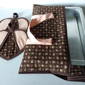 May include: A rectangular quilted casserole carrier with a brown floral pattern and light pink fabric handles, paired with two matching heart-shaped quilted pot holders, and a rectangular metal baking pan nestled inside the carrier.