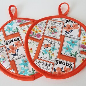 May include: Two round orange pot holders with a loop for hanging. The fabric features a white background with orange seed packet designs, showcasing various flowers. The edges are trimmed with orange piping.