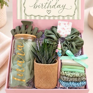 May include: A pink gift box containing a glass tumbler with a bamboo lid and gold text, a green hair claw clip, a succulent in a burlap pot, a floral-topped pen, a large olive green scrunchie, and a stack of smaller hair ties with a mint ribbon.