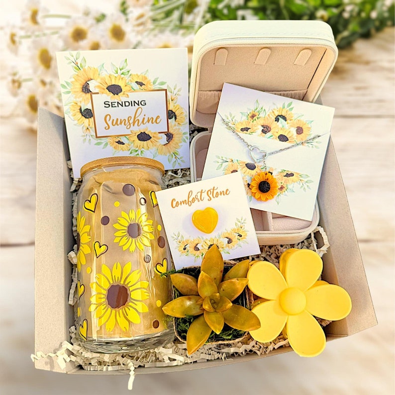 May include: A gift box filled with sunflower-themed items, including a glass jar with a sunflower design, a small succulent plant, a yellow flower-shaped silicone object, a card that says "Sending Sunshine", and a jewelry box with a sunflower necklace and a heart-shaped comfort stone.
