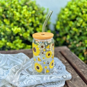 May include: A glass reusable water bottle with a bamboo lid and a straw. The bottle has a sunflower and heart pattern.
