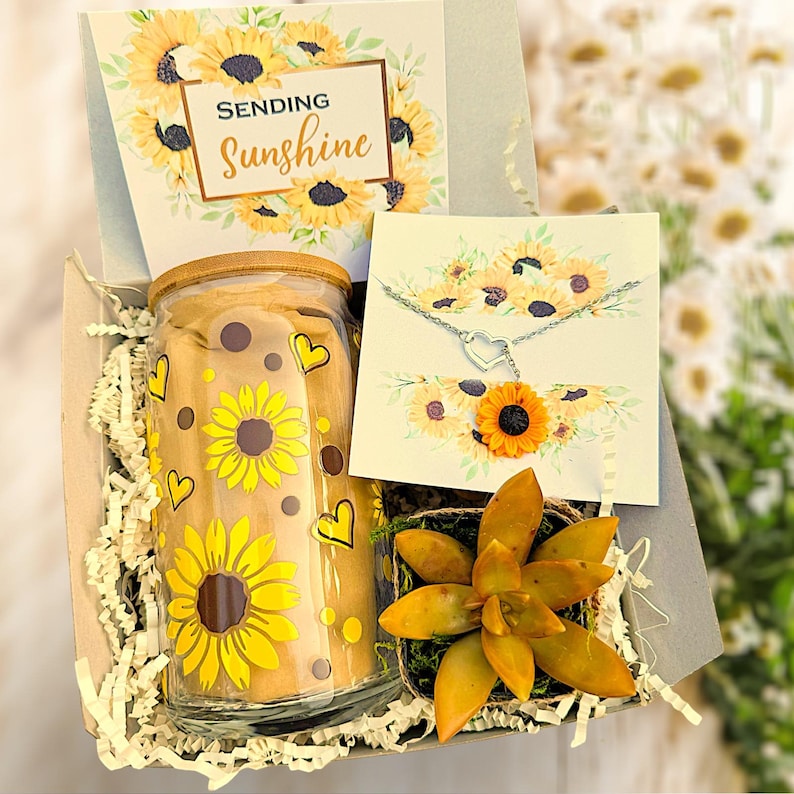 May include: A gift box filled with a sunflower patterned glass jar, a succulent plant, a card that says "Sending Sunshine", and a silver necklace with a heart pendant.