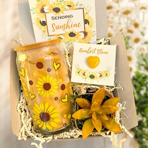 May include: A gift box filled with a glass jar with a sunflower design, a small succulent plant in a pot, a card that says "Sending Sunshine" and a card that says "Comfort Stone" with a yellow heart-shaped stone.