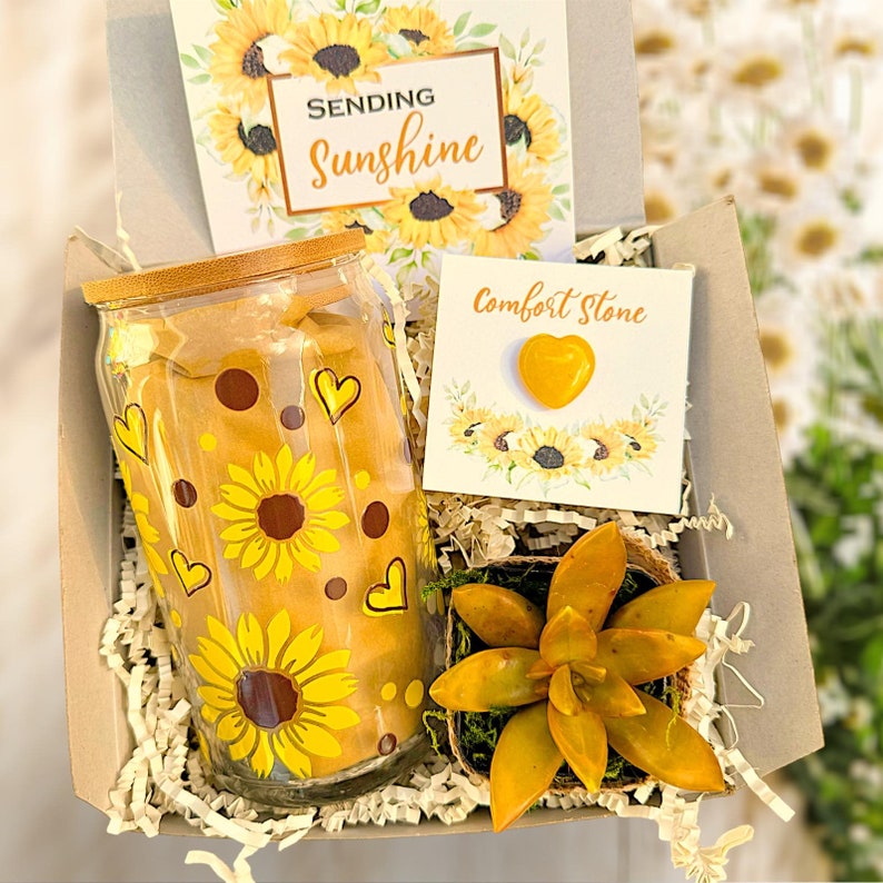 May include: A gift box filled with a glass jar with a sunflower design, a small succulent plant in a pot, a card that says "Sending Sunshine" and a card that says "Comfort Stone" with a yellow heart-shaped stone.