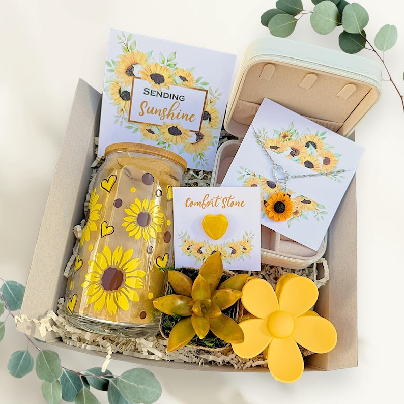 May include: A gift box filled with sunflower-themed items, including a glass jar with a sunflower design, a small succulent plant, a necklace with a sunflower pendant, a heart-shaped comfort stone, and a card that says "Sending Sunshine".