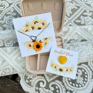 May include: A sunflower necklace and a heart-shaped comfort stone in a jewelry box with a sunflower design. The necklace has a silver chain and a sunflower charm. The comfort stone is yellow and heart-shaped.