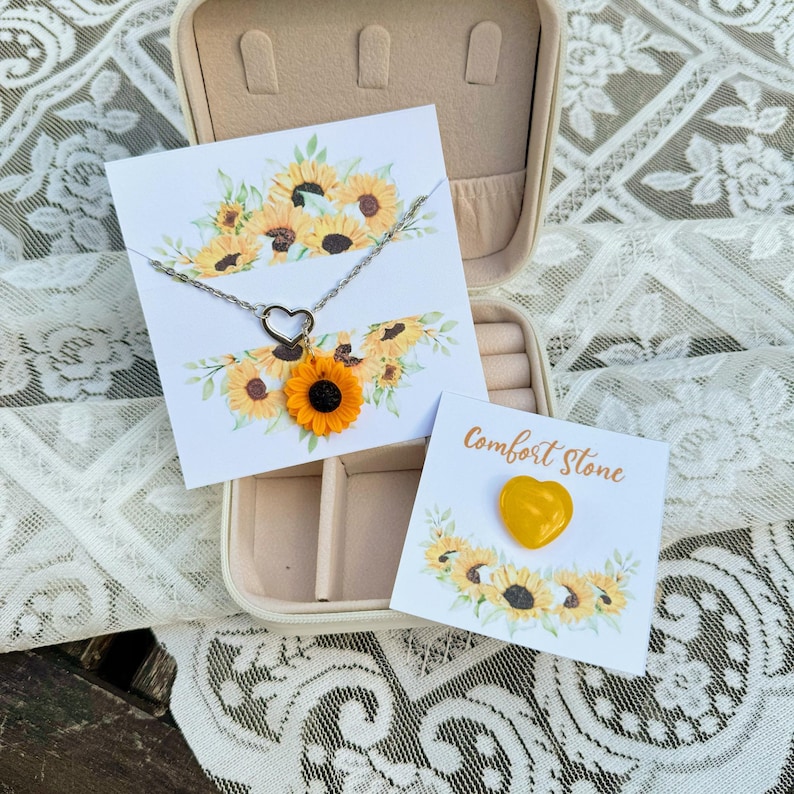 May include: A sunflower necklace and a heart-shaped comfort stone in a jewelry box with a sunflower design. The necklace has a silver chain and a sunflower charm. The comfort stone is yellow and heart-shaped.