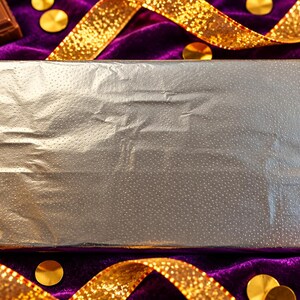 May include: A rectangular chocolate bar wrapped in smooth, reflective silver foil. The metallic packaging has a subtle, fine-textured surface pattern and is positioned horizontally, centered in the frame.