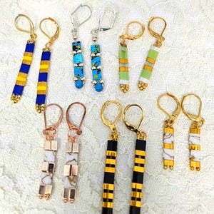 Pode incluir: Uma coleção de brincos feitos à mão com fechos de alavanca. Os brincos apresentam contas coloridas em azul, amarelo, verde, branco e preto. Alguns brincos têm um padrão listrado. Estilos: dourado, prateado e cobre.