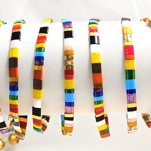 Puede incluir: Una colección de pulseras de cuentas coloridas. Cada pulsera presenta un diseño de cuentas rectangulares en una variedad de colores, incluyendo rojo, amarillo, azul, verde y dorado. Las pulseras se exhiben sobre una superficie blanca.