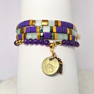 Peut inclure: Plusieurs bracelets superposés, composés de perles violettes, dorées et vert clair. L'un des bracelets est orné d'un charm avec un pendentif rond doré et une feuille. Les bracelets sont présentés sur une surface blanche.