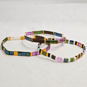May include: Three colorful beaded bracelets. Each bracelet features small, rectangular beads in various colors, including green, blue, purple, and gold. The bracelets have gold-colored accents and are displayed on a white surface.