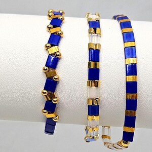 May include: Three beaded bracelets. One features a zig-zag pattern of navy blue and gold beads. Another has a vertical design of navy blue, gold, and clear beads. The third bracelet has a straight design of navy blue and gold beads. Jewelry.