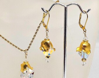 May include: A gold-toned jewelry set featuring a necklace and earrings. Each piece has a gold-colored flower-shaped top and a clear, faceted crystal pendant. The earrings hang from gold-colored lever-back closures.