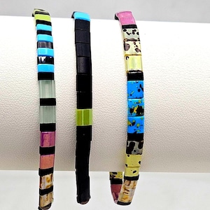May include: Three colorful beaded bracelets. One bracelet features black, blue, and green beads. Another has black and green beads. The third bracelet has pink, yellow, and blue beads. Each bracelet is made of rectangular beads.