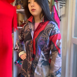 Könnte beinhalten: Eine gemusterte Jacke in Rot-, Braun- und Lilatönen. Die Jacke hat einen Kragen, lange Ärmel mit gestreiften Manschetten und wird über einem rosa Oberteil und einer blauen Jeans getragen. Die Jacke hat eine Knopfleiste.